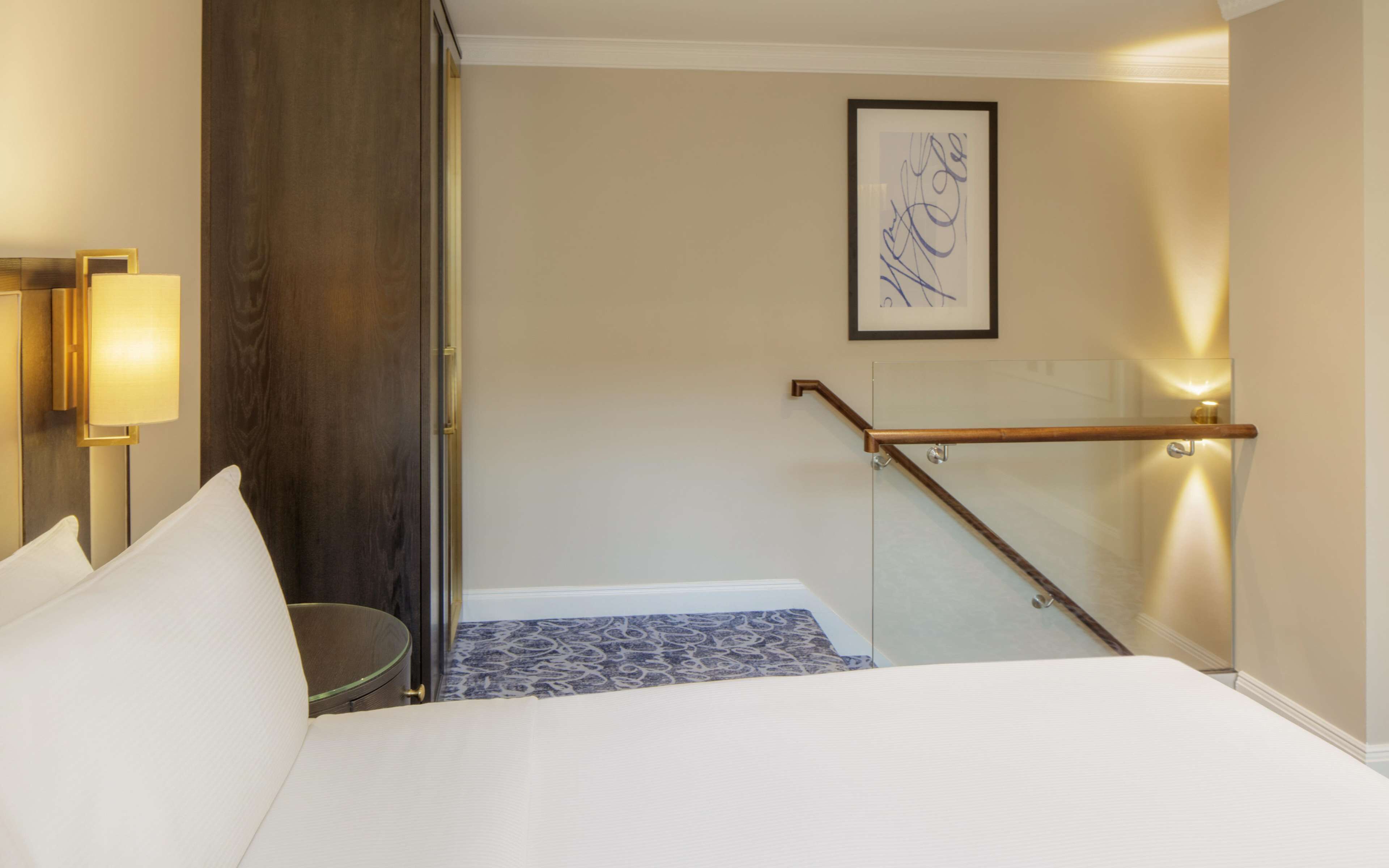 A hotel room with a bed, a nightstand, a lamp, a wooden door, a staircase, and a framed artwork on the wall.