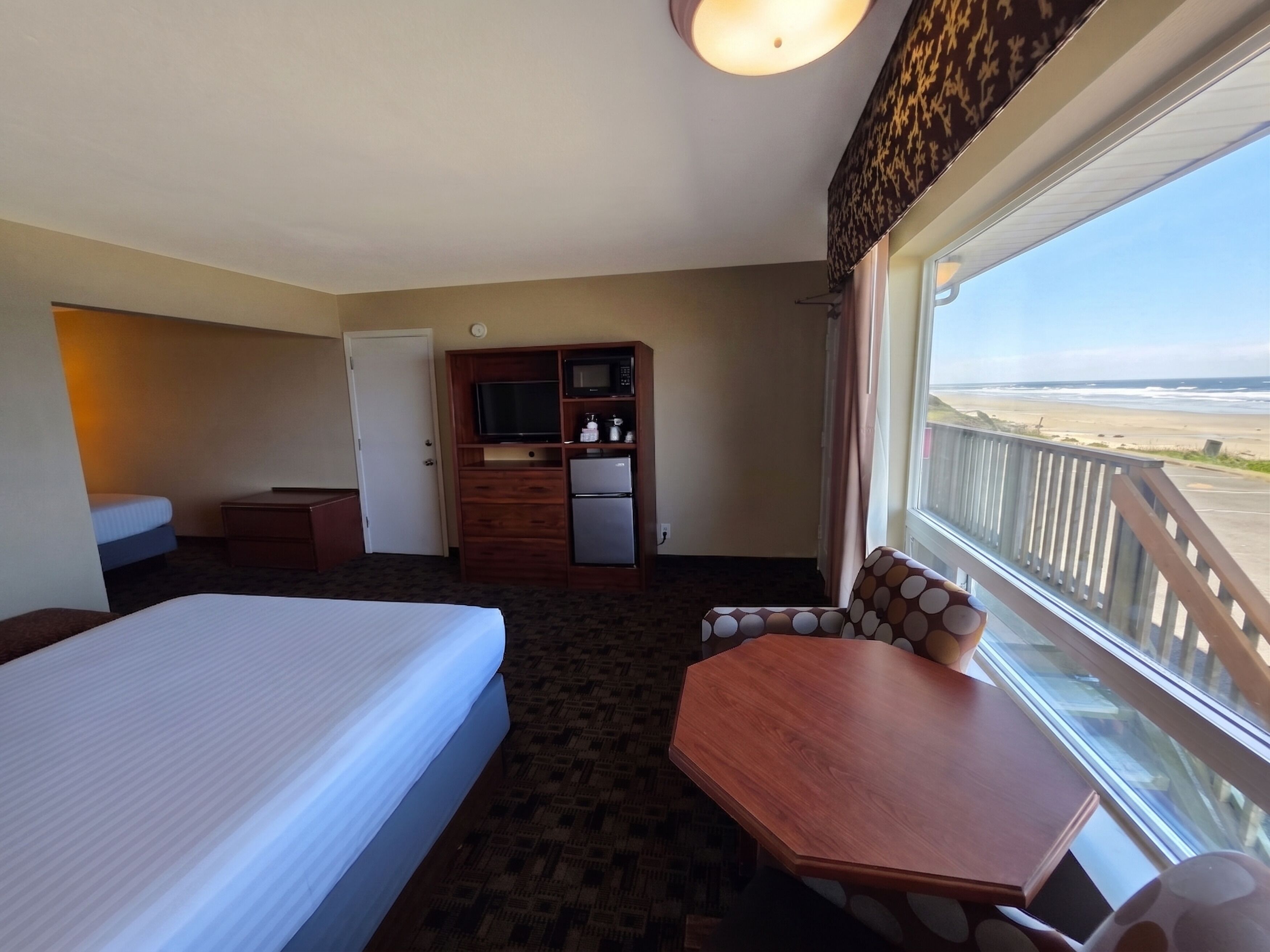 A hotel room with a bed, a desk, a television, a refrigerator, and a view of the beach.