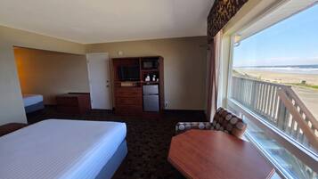 A hotel room with a bed, a desk, a television, a refrigerator, and a view of the beach.