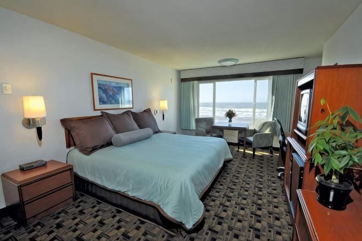 A hotel room with a bed, bedside lamps, a desk, a chair, a television, and a window with a view of the ocean.