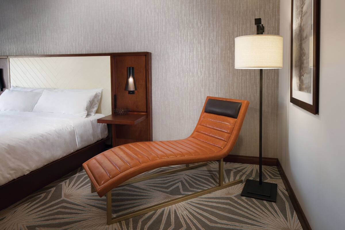 Ein Hotelzimmer mit Bett, Sessel, Nachttisch, Lampe und einem gerahmten Bild an der Wand.