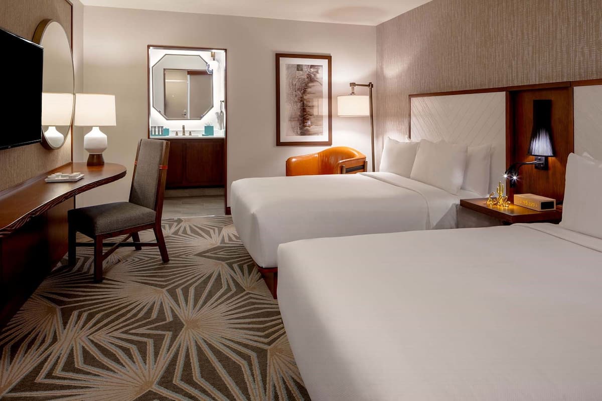 Ein Hotelzimmer mit zwei Betten, einem Schreibtisch mit Stuhl, einem Fernseher, einem großen Spiegel und einem gerahmten Kunstwerk an der Wand.
