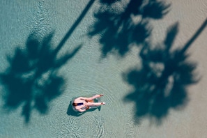 An aerial view of a person floating on a clear body of water, surrounded by shadows of tree branches.