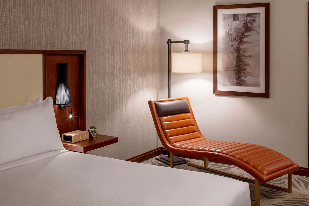 Ein Hotelzimmer mit Bett, Sessel, Nachttisch mit Lampe, gerahmtem Kunstwerk und einer Uhr.