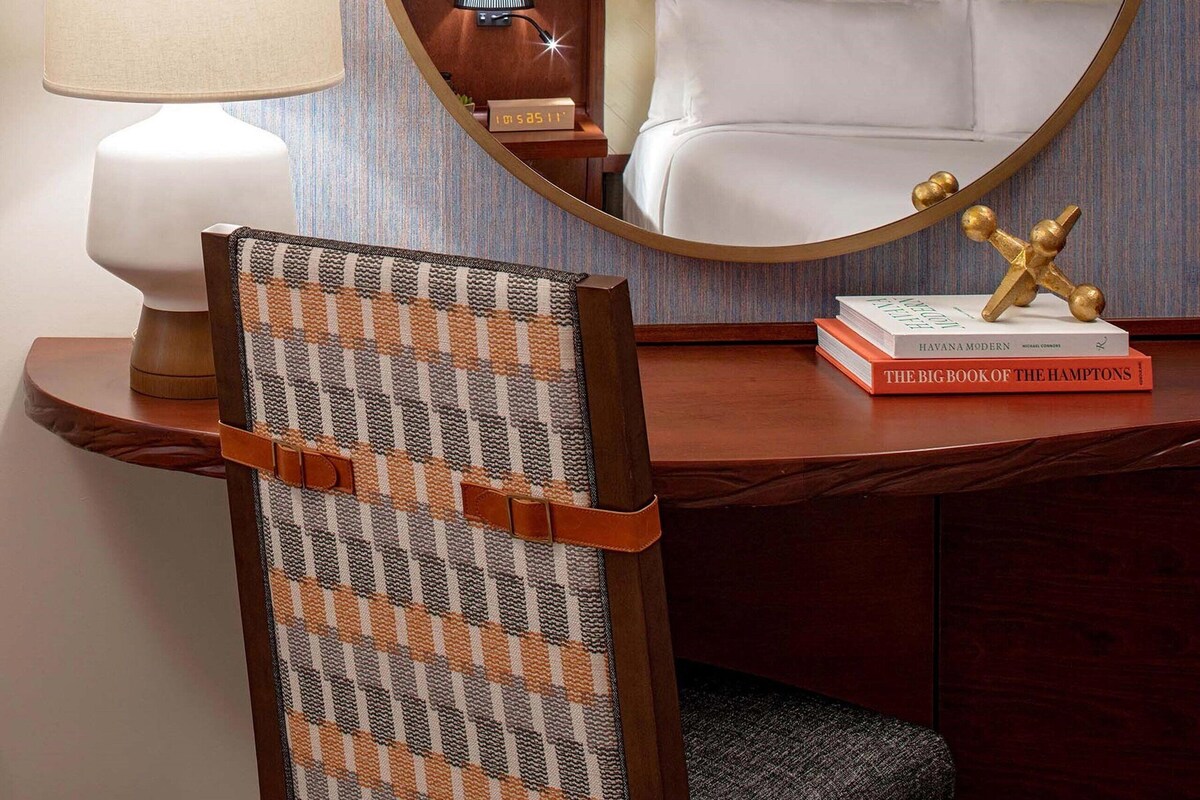 A hotel room with a round mirror, a wooden desk, a chair, a lamp, a bedside table with a lamp, a bed, and a book titled "The Big Book of the Hamptons".