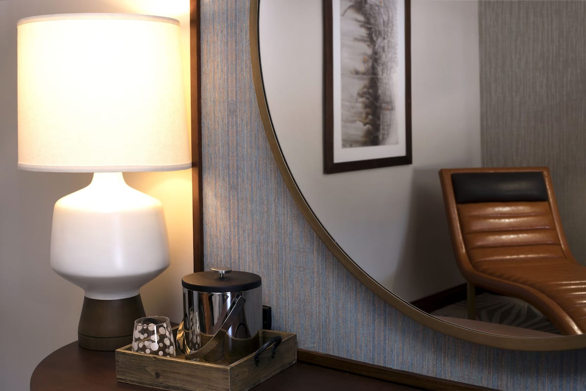 Ein Hotelzimmer mit einem runden Tisch, einer Lampe, einem Spiegel, einem gerahmten Bild an der Wand und einem Ledersessel.