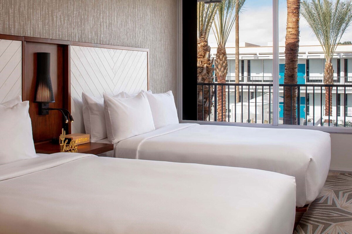 A hotel room with two beds, a nightstand with a lamp, and a view of palm trees and a building outside.