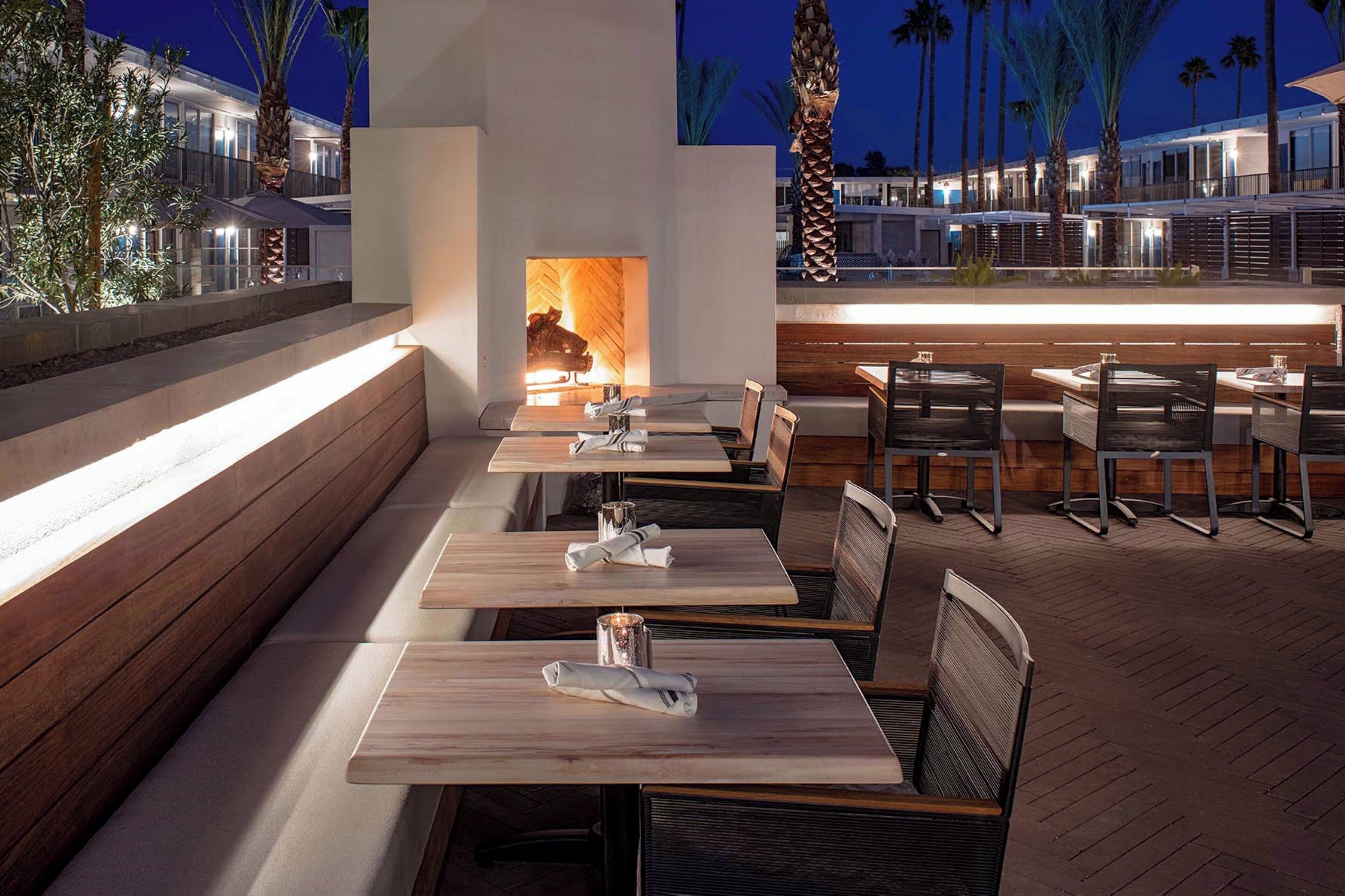 An outdoor dining area with wooden tables and benches, a fire feature, and a view of a modern building in the background.