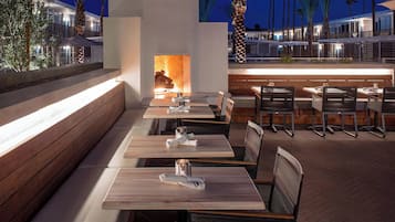 An outdoor dining area with wooden tables and benches, a fire feature, and a view of a modern building in the background.