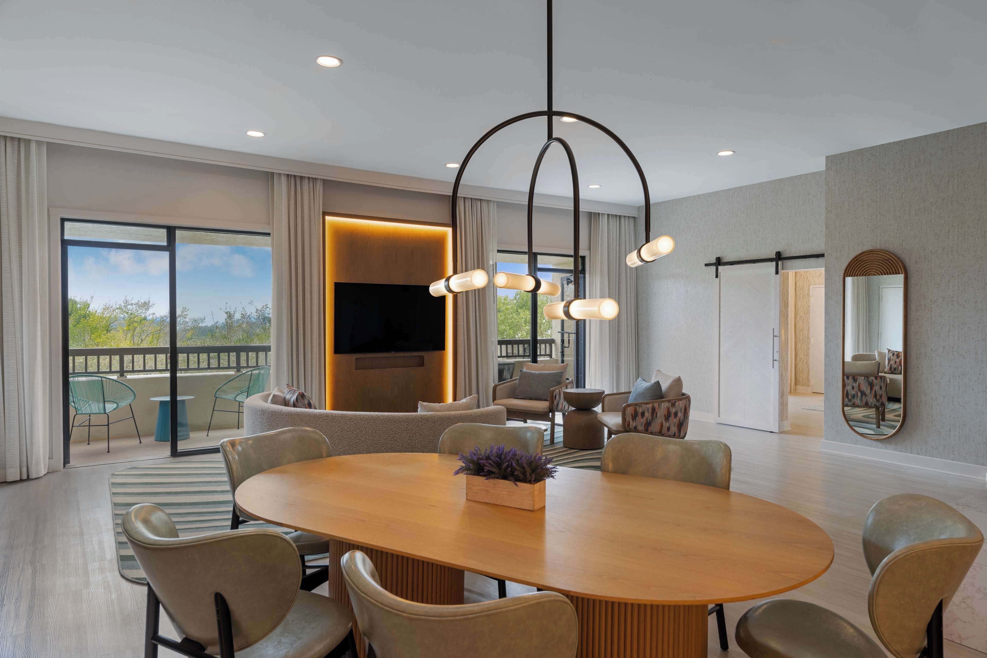 A spacious living room with a large round table, comfortable seating, a modern chandelier, and a large window offering a view of the outdoors.