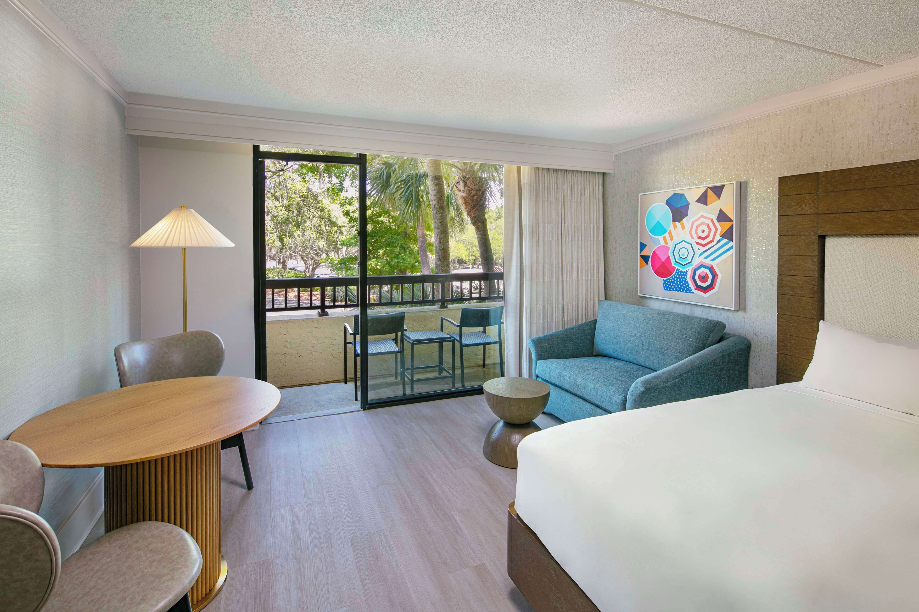 A hotel room with a bed, a sofa, a table, and a chair. There is a balcony with a view of trees and a railing.