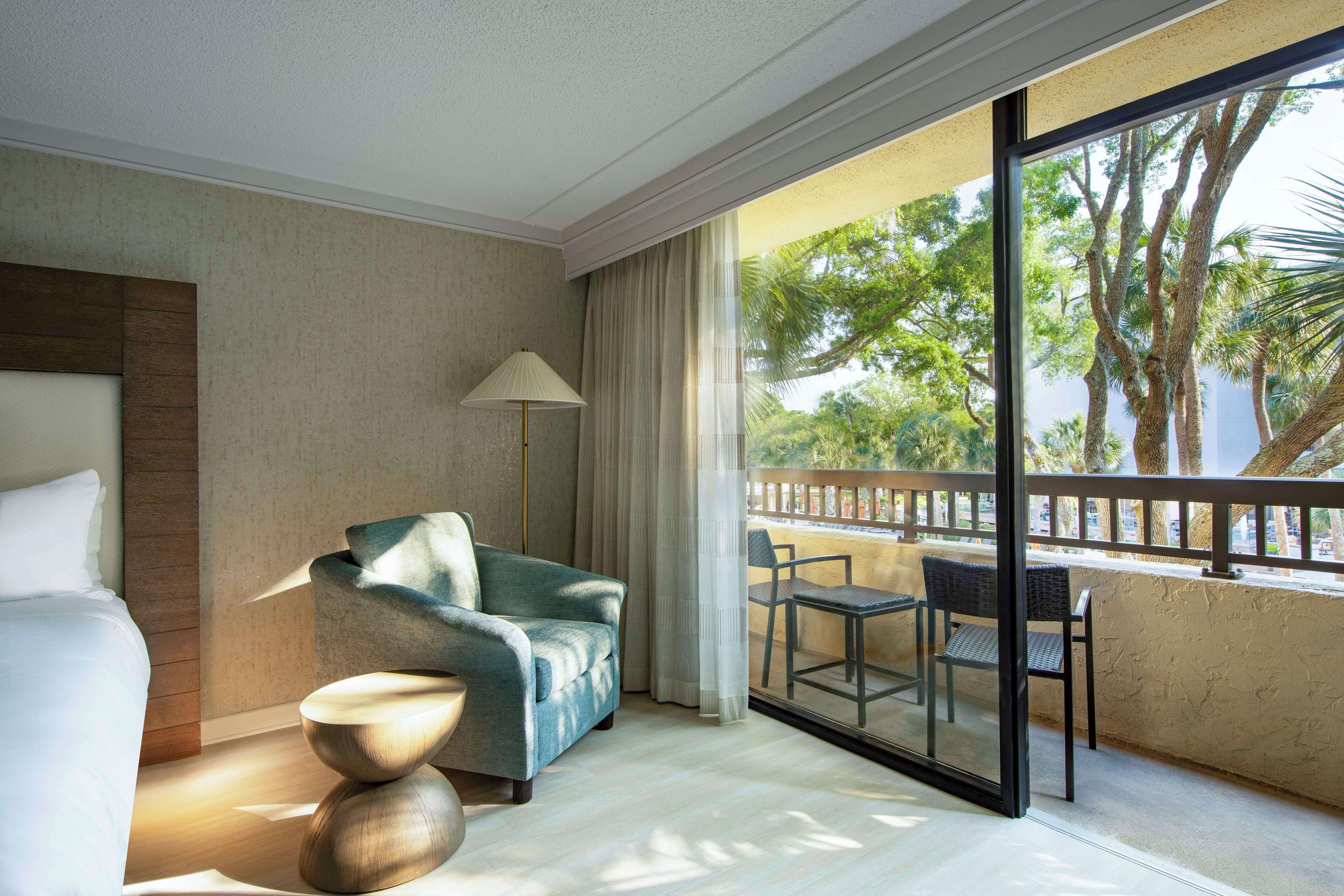 A hotel room with a bed, a sofa, a table, and a balcony with a view of trees.