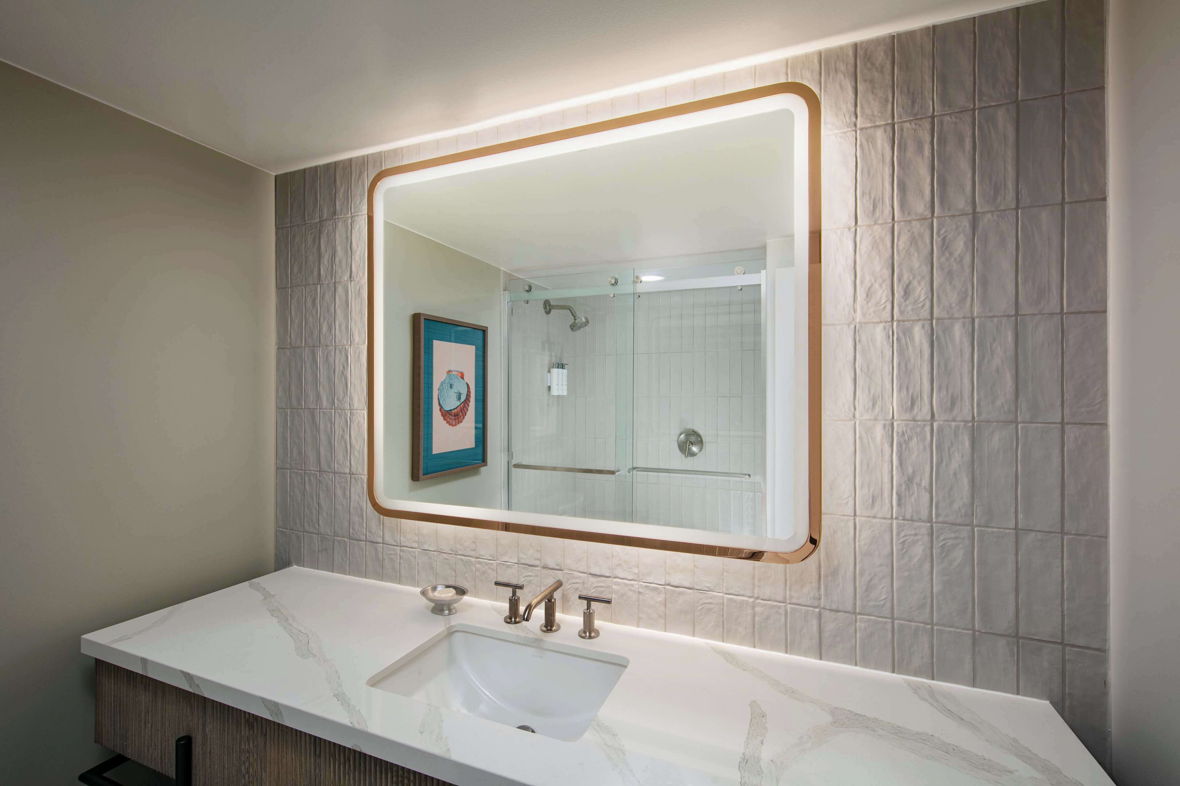 Une salle de bain avec un lavabo en marbre, un tableau encadré au mur et une douche visible dans le reflet du miroir.
