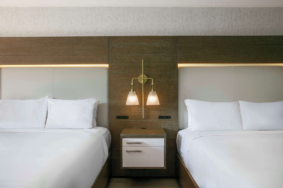 Habitación de hotel con dos camas, una mesita de noche con cajón y una lámpara fijada a la pared.