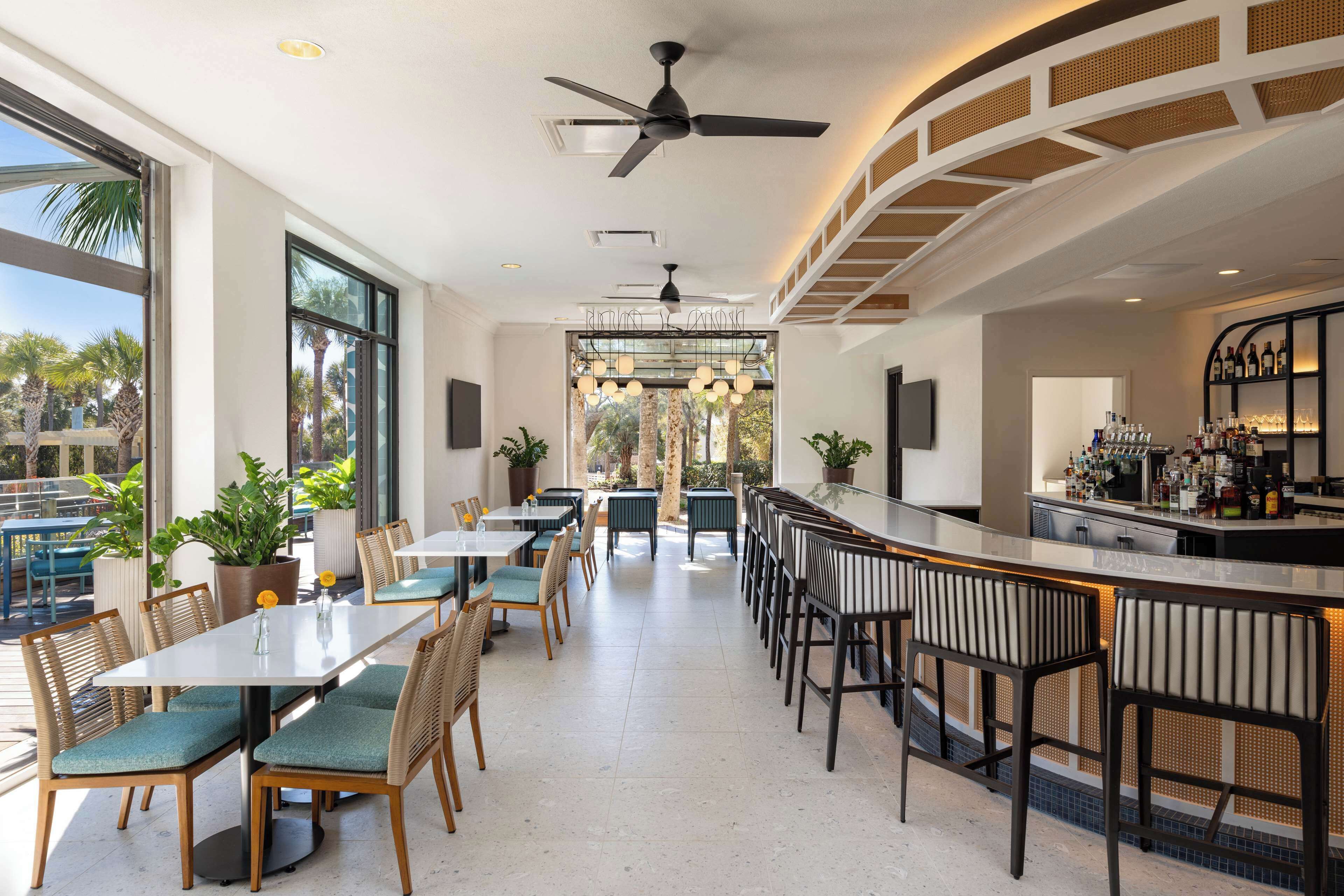 Un restaurante moderno con zona de bar, ventanas amplias y plantas de interior.