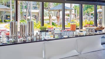 A buffet setup with stainless steel dispensers, a variety of food items, and a view of a tropical outdoor area through large windows.