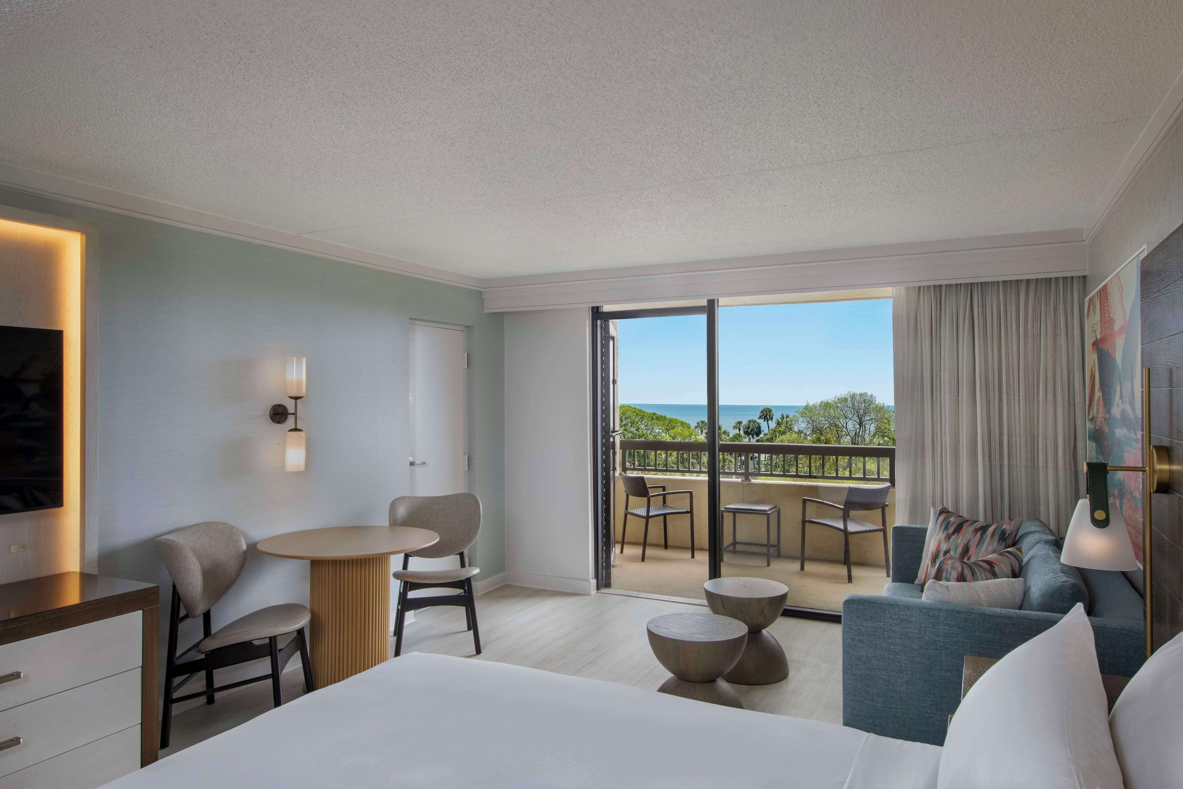 Quarto de hotel com cama, escrivaninha, cadeira, mesa pequena, sofá e varanda com vista para o mar.