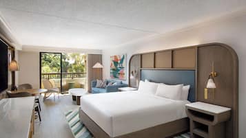 A hotel room with a large bed, a desk, a chair, a sofa, and a balcony with a view of greenery.