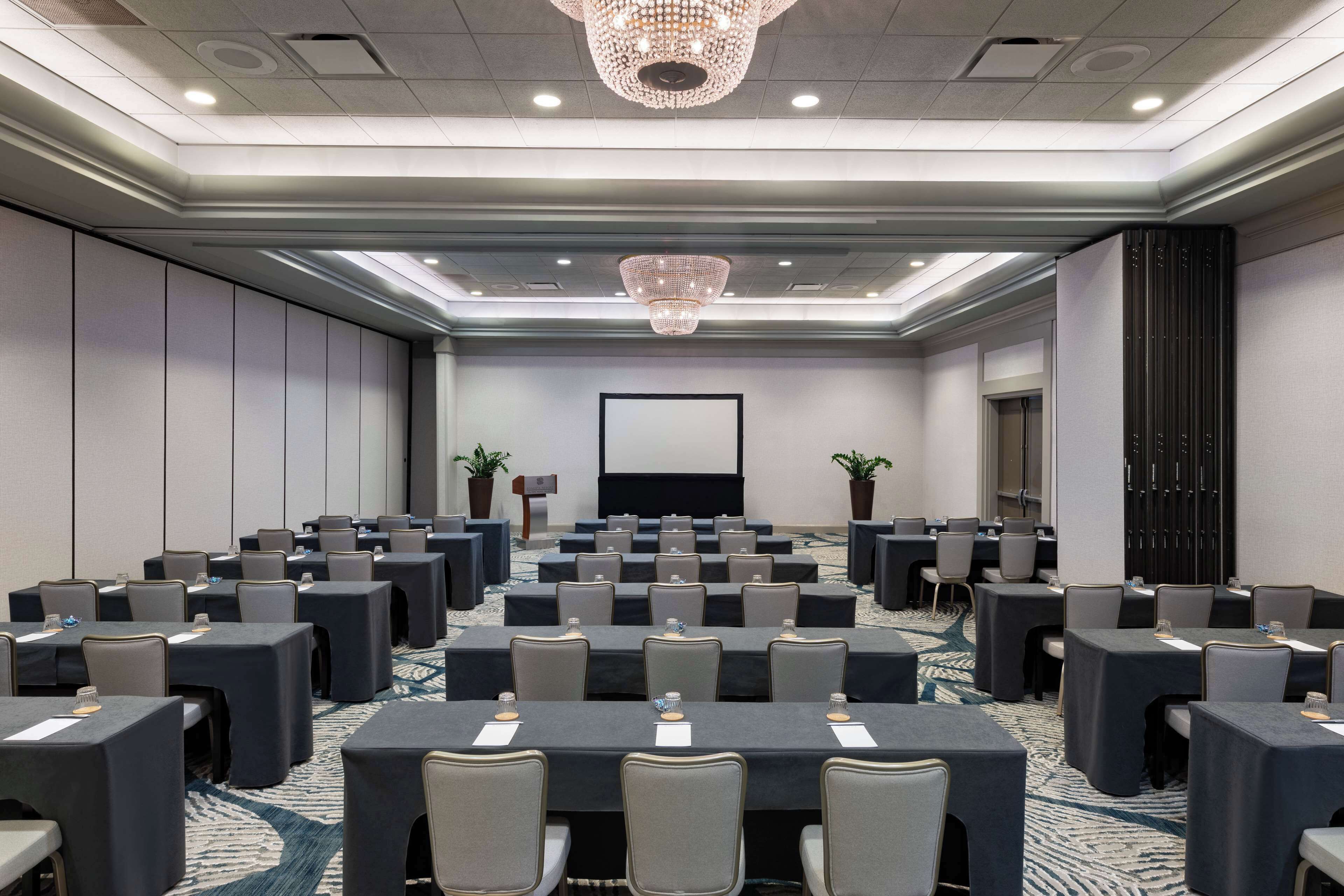 A conference room with multiple tables and chairs, a large screen, and a chandelier.