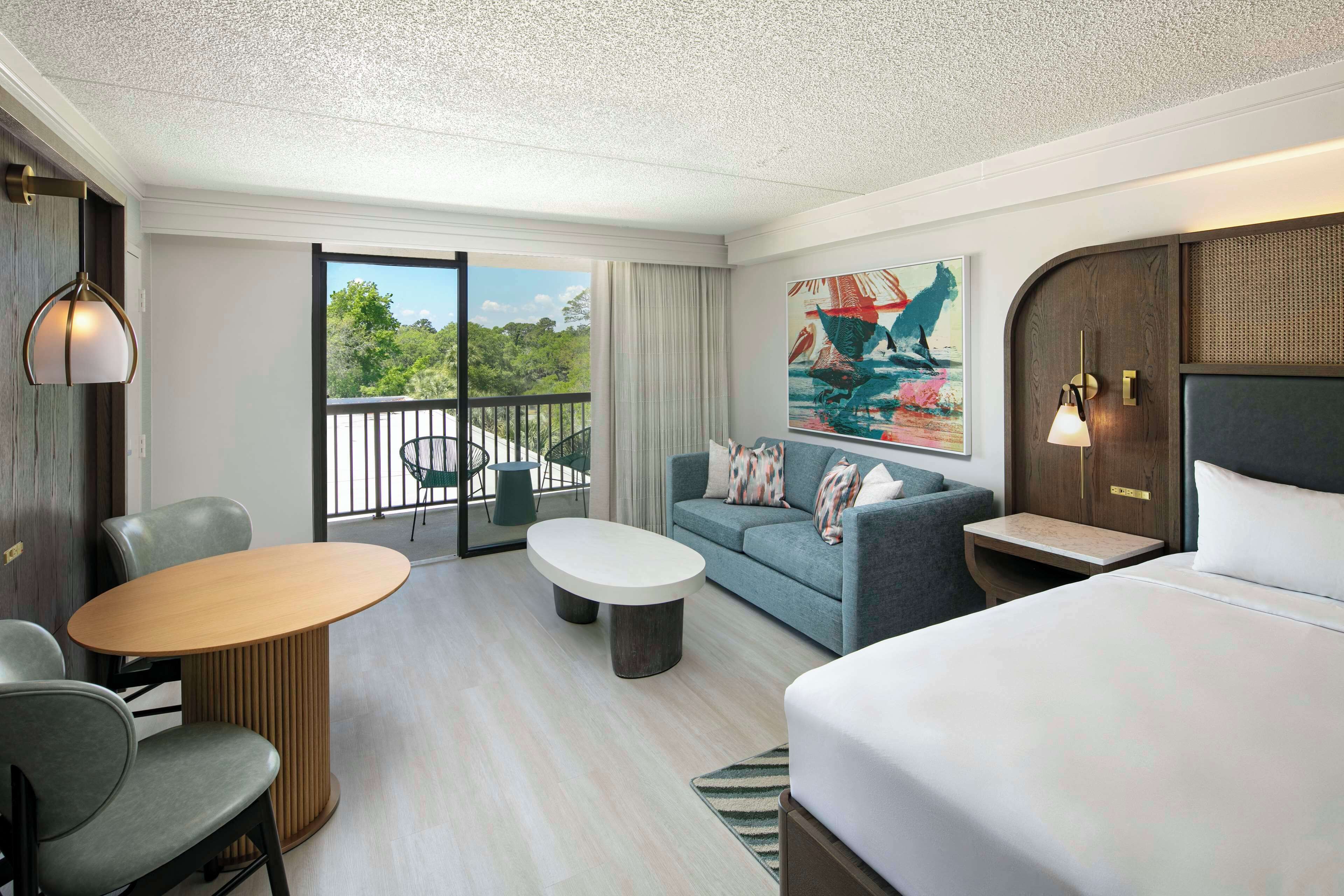 A hotel room with a bed, a sofa, a dining table, and a balcony with a view of greenery.