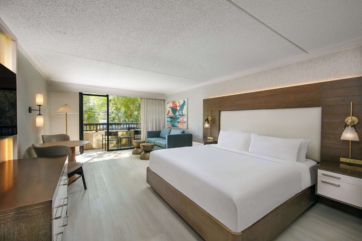 Habitación de hotel con una cama grande, un escritorio, una silla, un sofá y un balcón con vistas a zonas verdes.