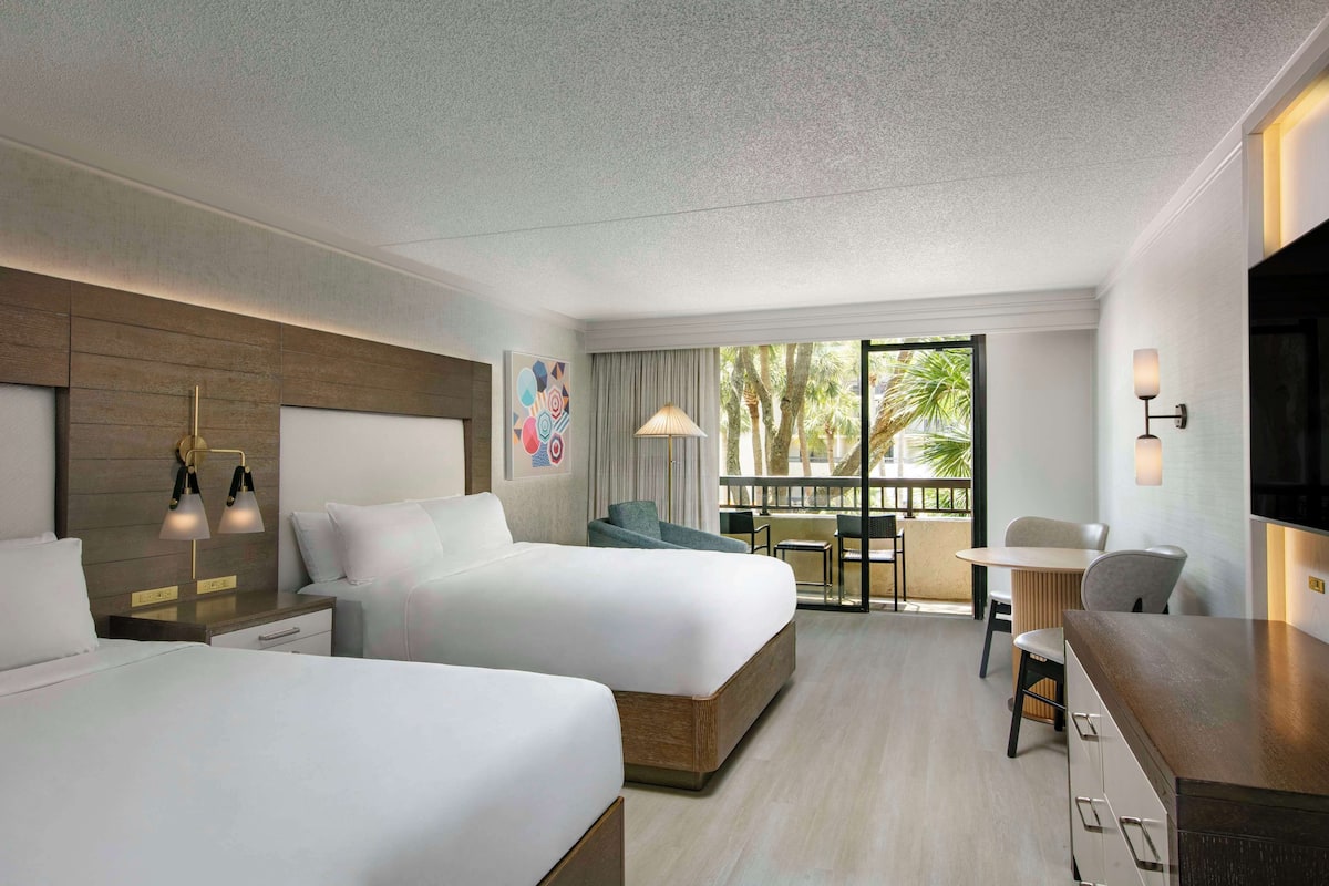 Una habitación de hotel moderna con dos camas, un escritorio, una silla y un balcón con vistas a palmeras.