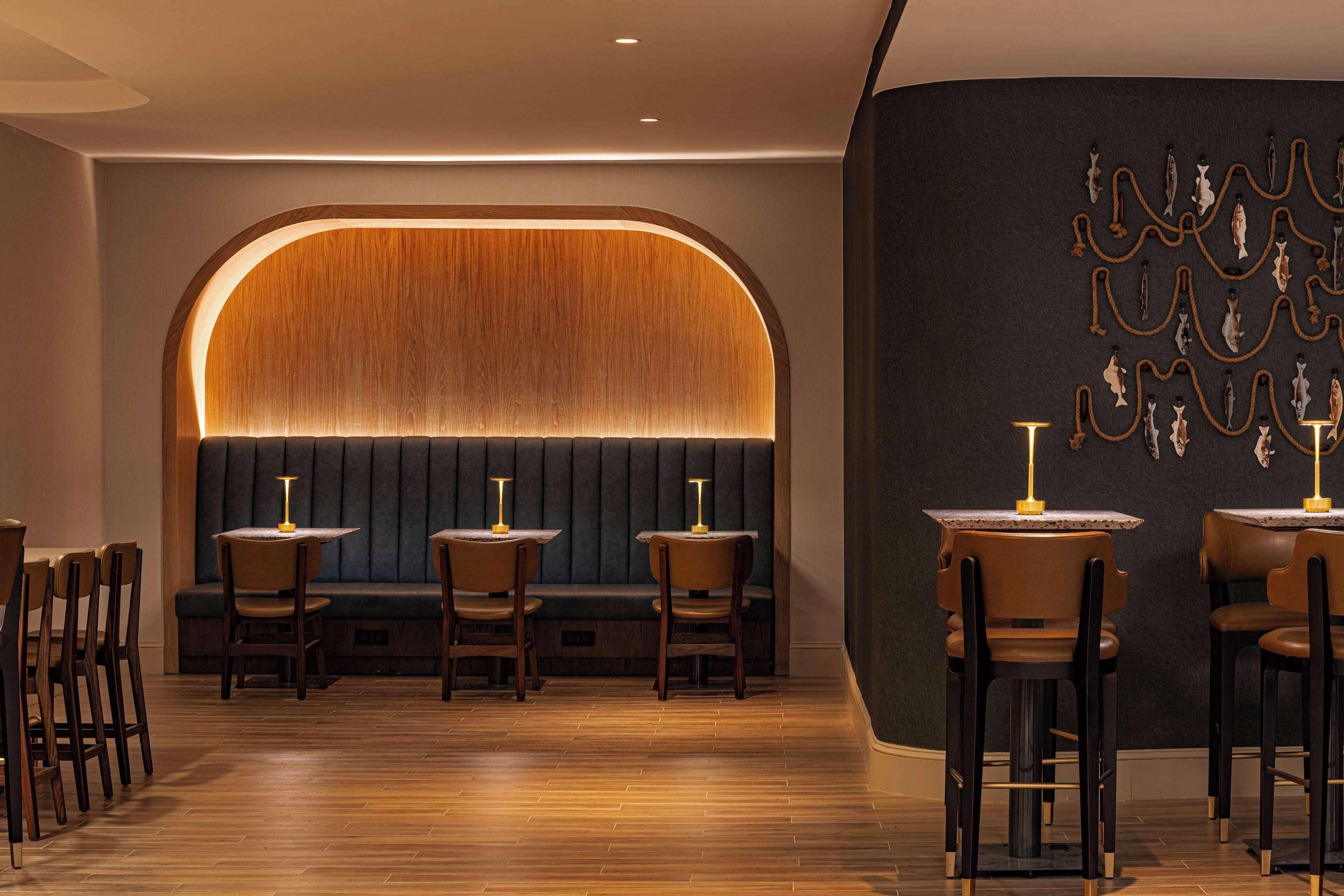 A restaurant with a wooden bench seating area, tables with lamps, and a dark wall with hanging decorations.