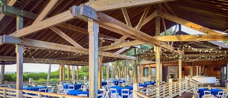 Un área de comedor al aire libre cubierta con vigas de madera, barandillas blancas y manteles azules.