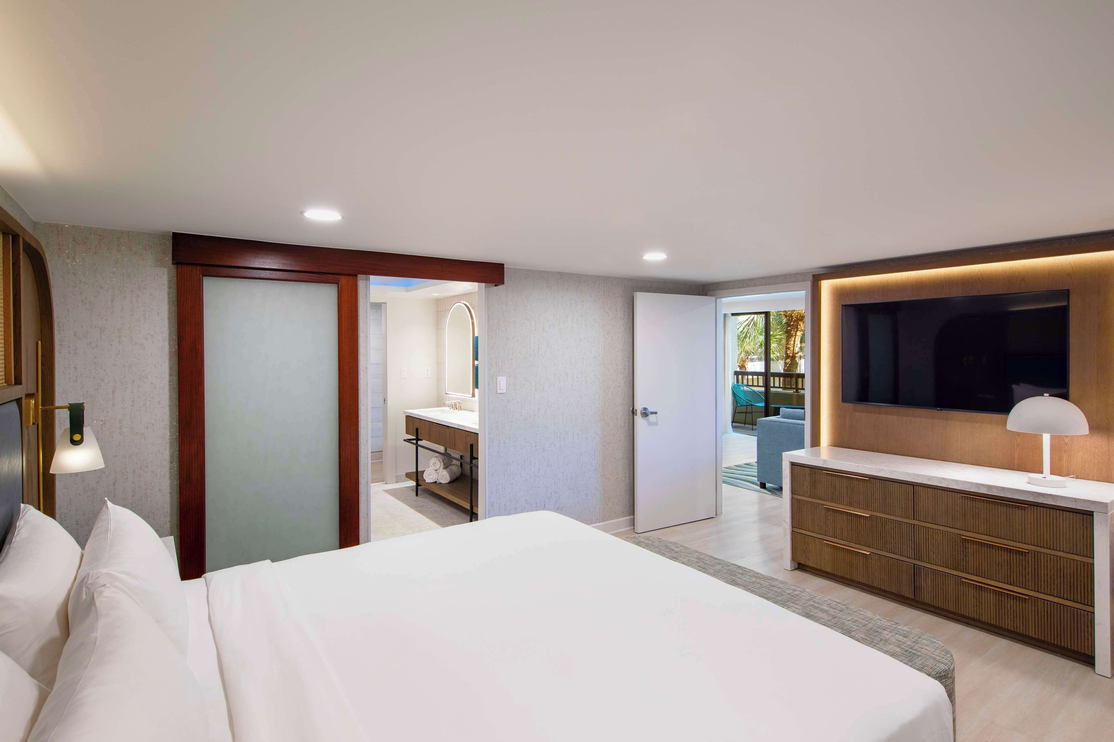 A modern hotel room with a large bed, a TV, a desk, and a bathroom visible through an open door.