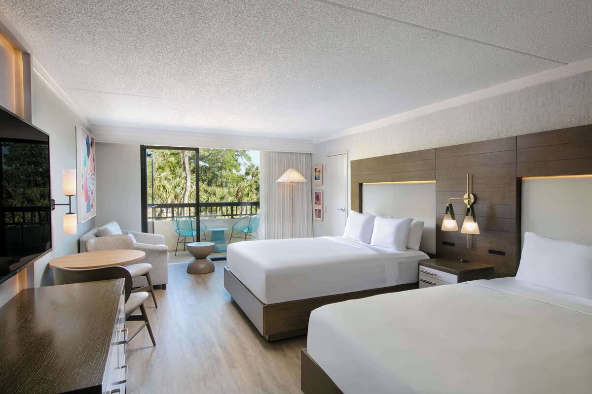 Habitación de hotel moderna con dos camas, zona de comedor y balcón con vistas a zonas verdes.