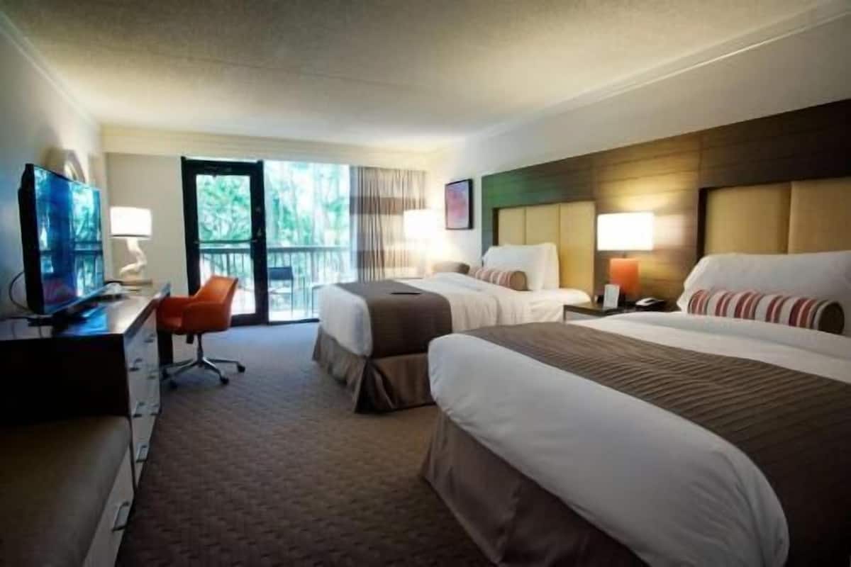 Ein Hotelzimmer mit zwei Betten, einem Schreibtisch mit Computer, einem Stuhl und einem Fenster mit Blick auf Bäume.
