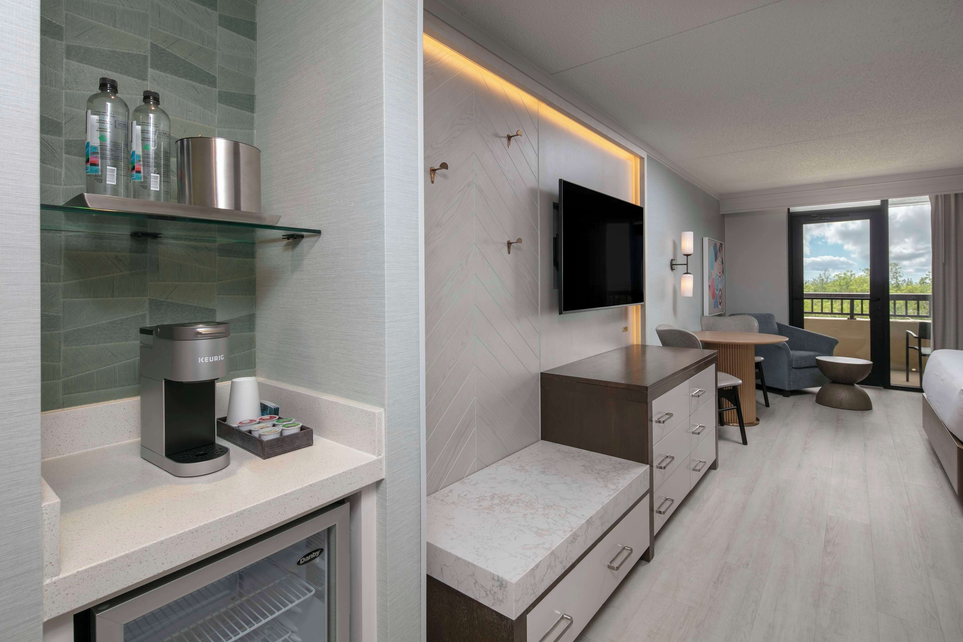 Quarto de hotel moderno com cozinha compacta, televisão de tela plana na parede, área de estar com mesa e cadeiras, e varanda com vista.