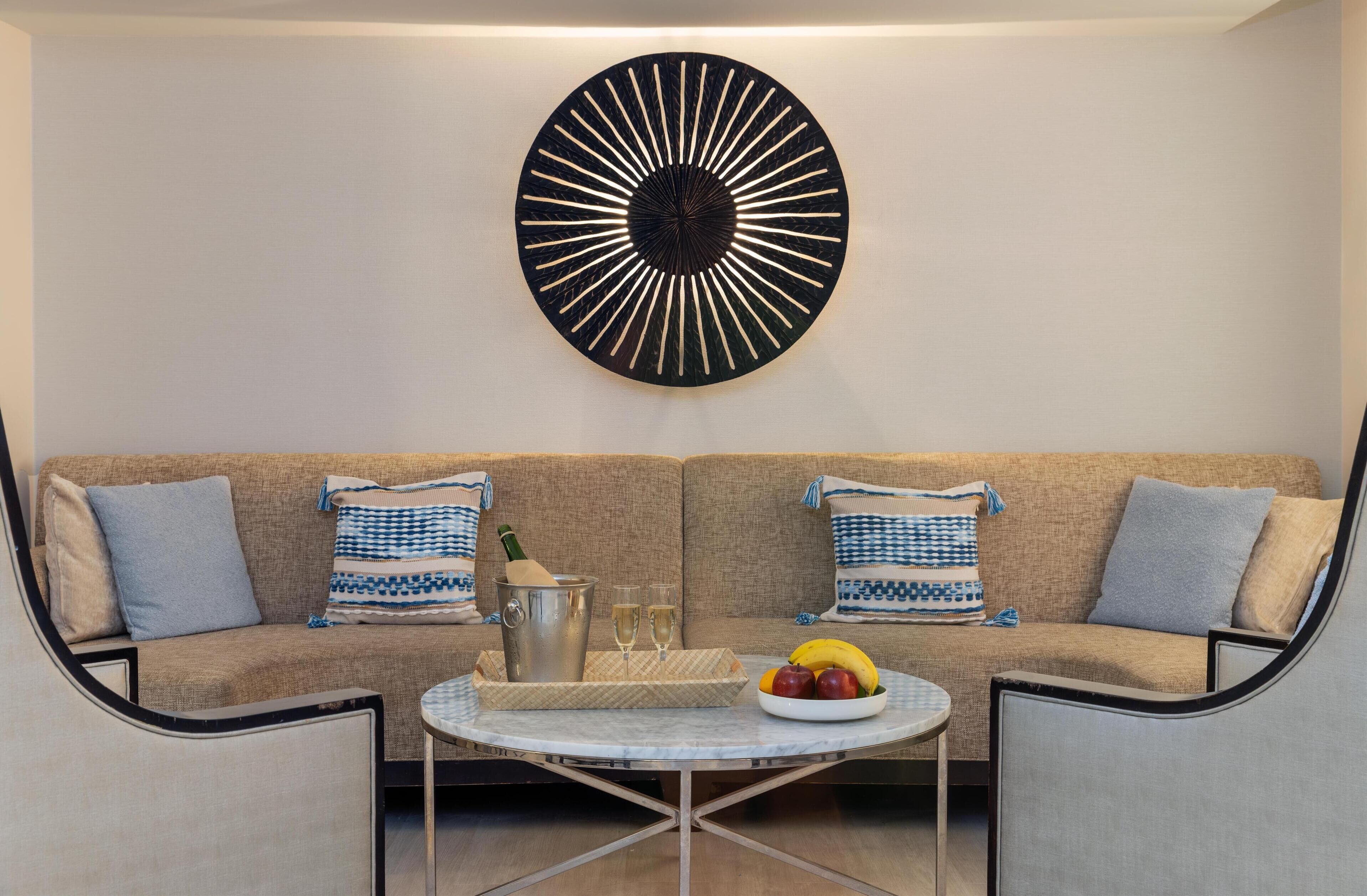 Un soggiorno moderno con un divano beige, due cuscini decorativi a motivi, un tavolino rotondo con una ciotola di frutta e un elemento decorativo a parete.