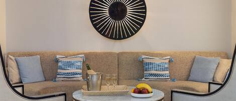 A modern living room with a beige sofa, two patterned throw pillows, a round coffee table with a fruit bowl, and a decorative wall piece.