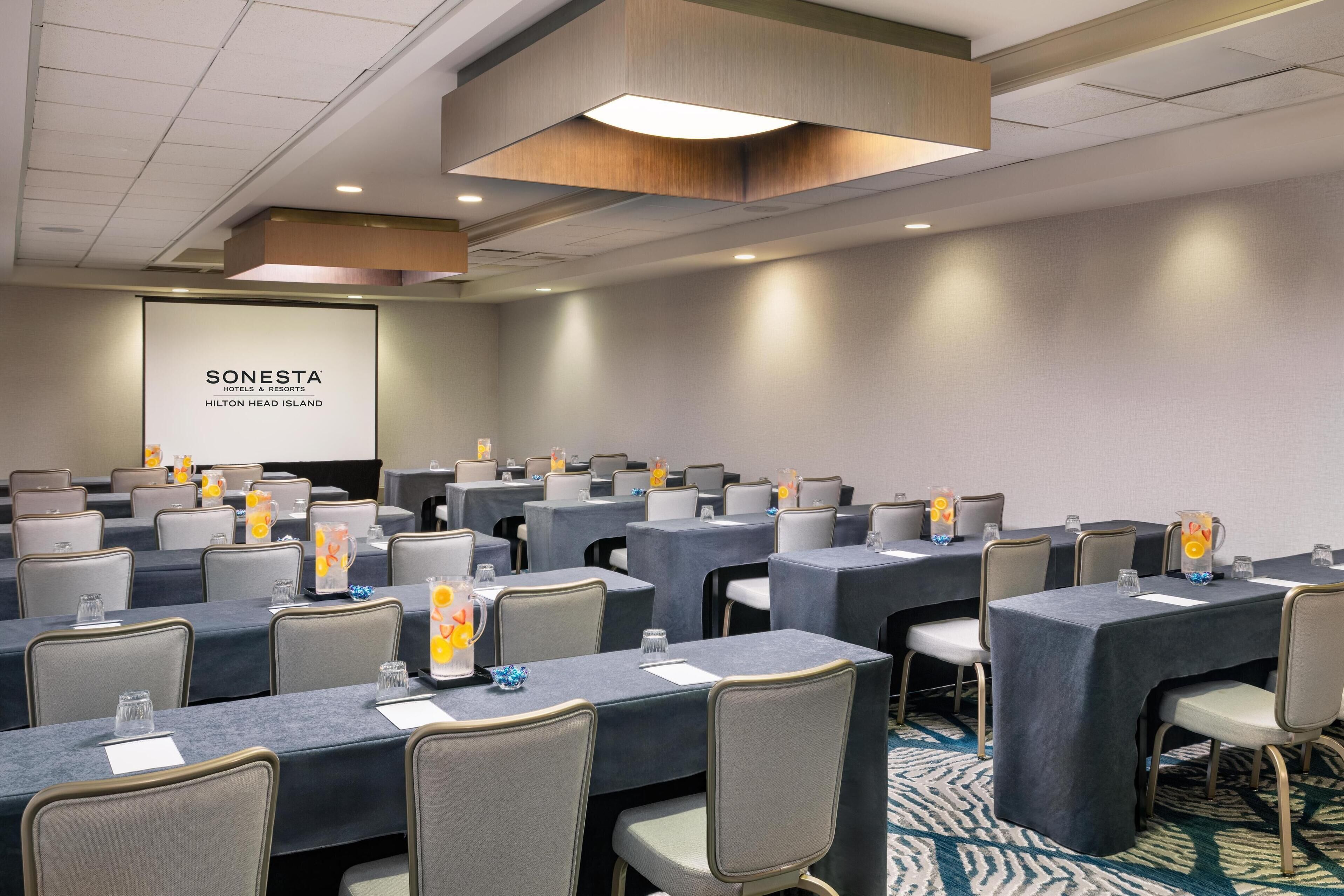 A conference room with multiple tables and chairs, a projector screen displaying 'SONESTA HOTELS & RESORTS HILTON HEAD ISLAND', and a modern ceiling design.
