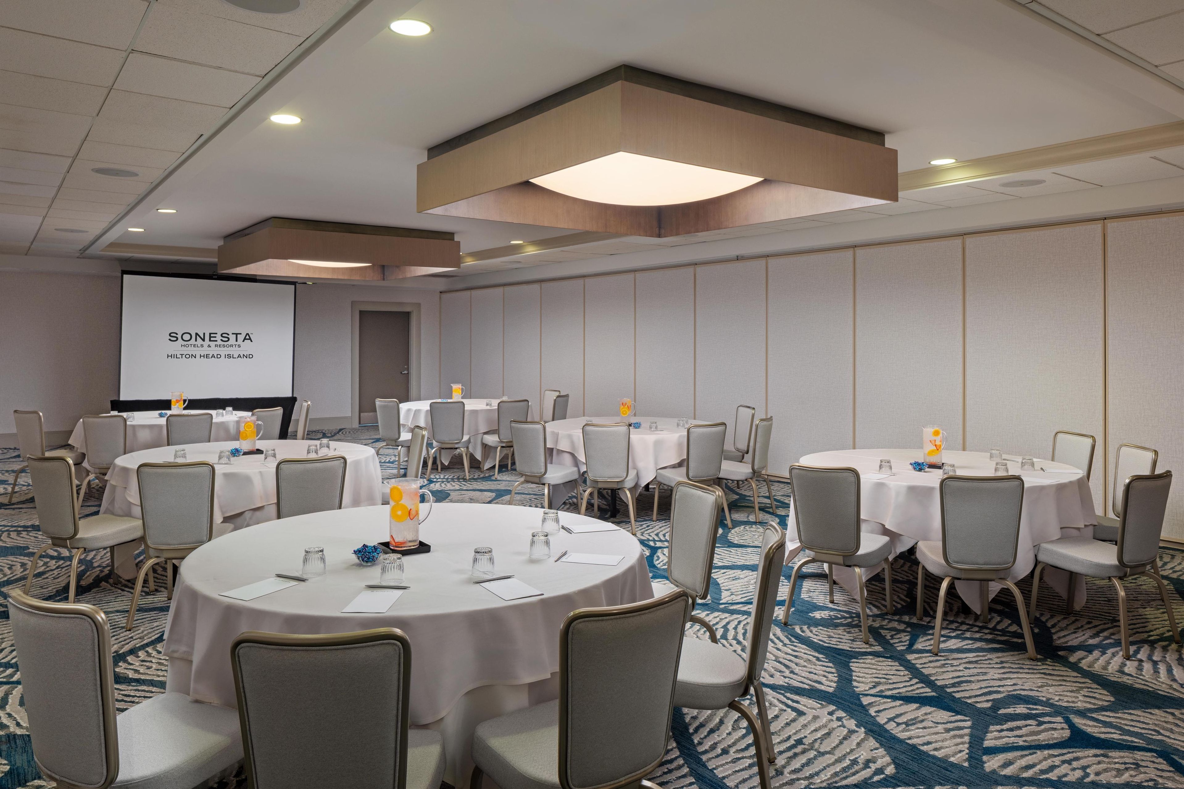 A conference room with multiple round tables, each set with chairs and white tablecloths. A projector screen displays the text "SONESTA HOTEL & RESORTS HILTON HEAD ISLAND." The room features a patterned carpet and recessed ceiling lighting.