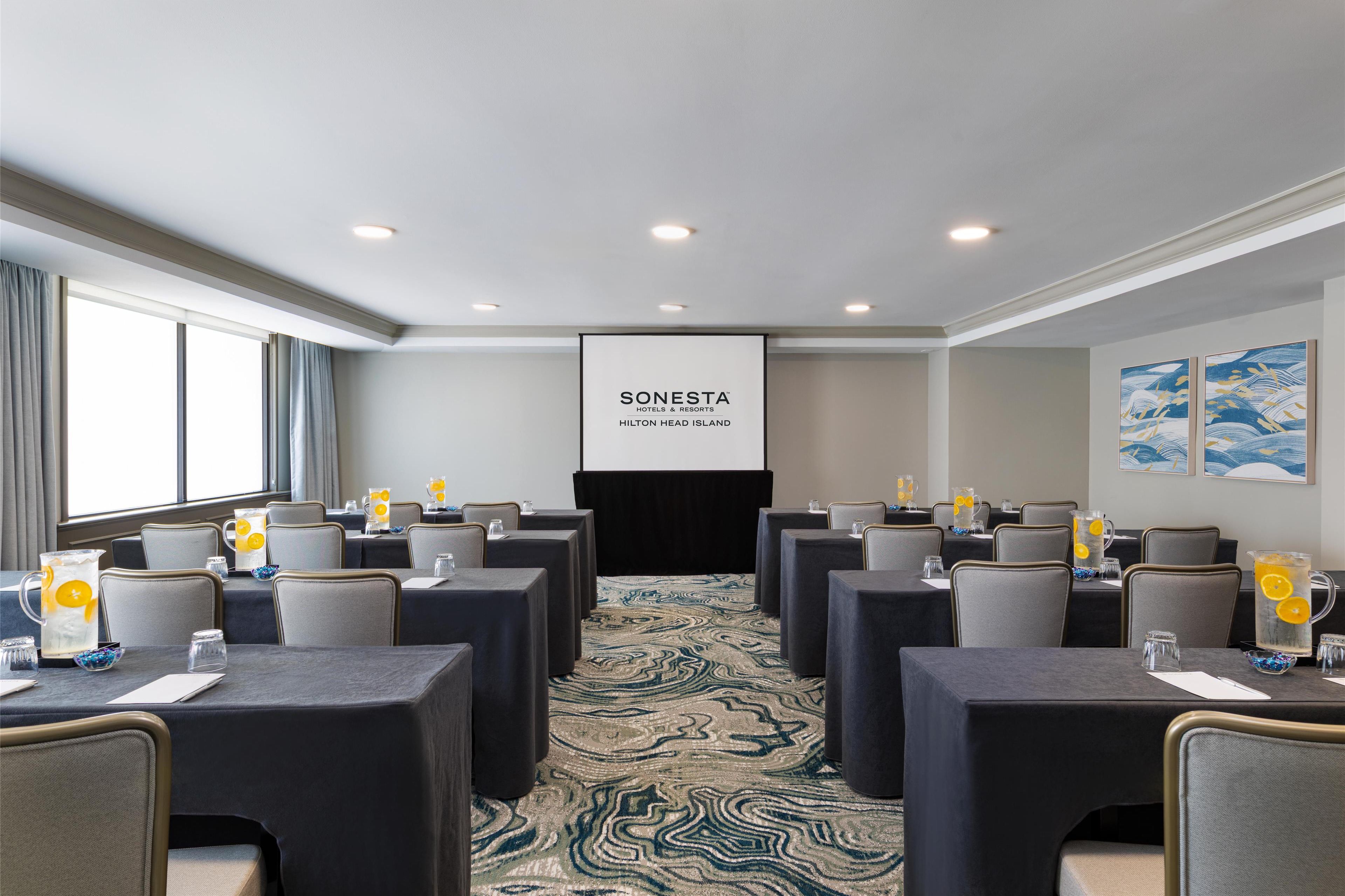 A conference room with tables, chairs, and a projector screen displaying "SONESTA HILTON HEAD ISLAND".