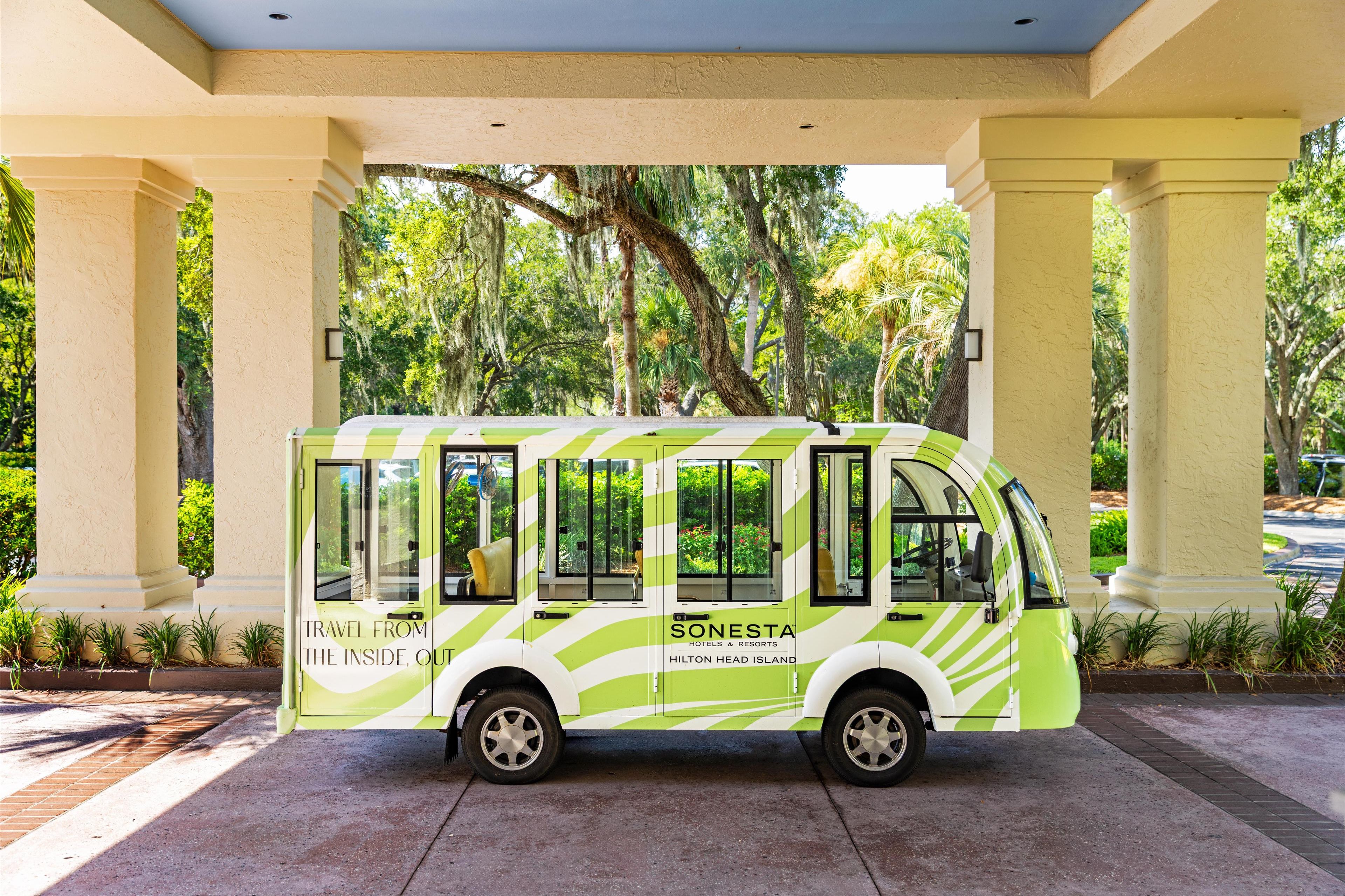 A green and white shuttle bus parked under a covered area with columns, surrounded by lush greenery.