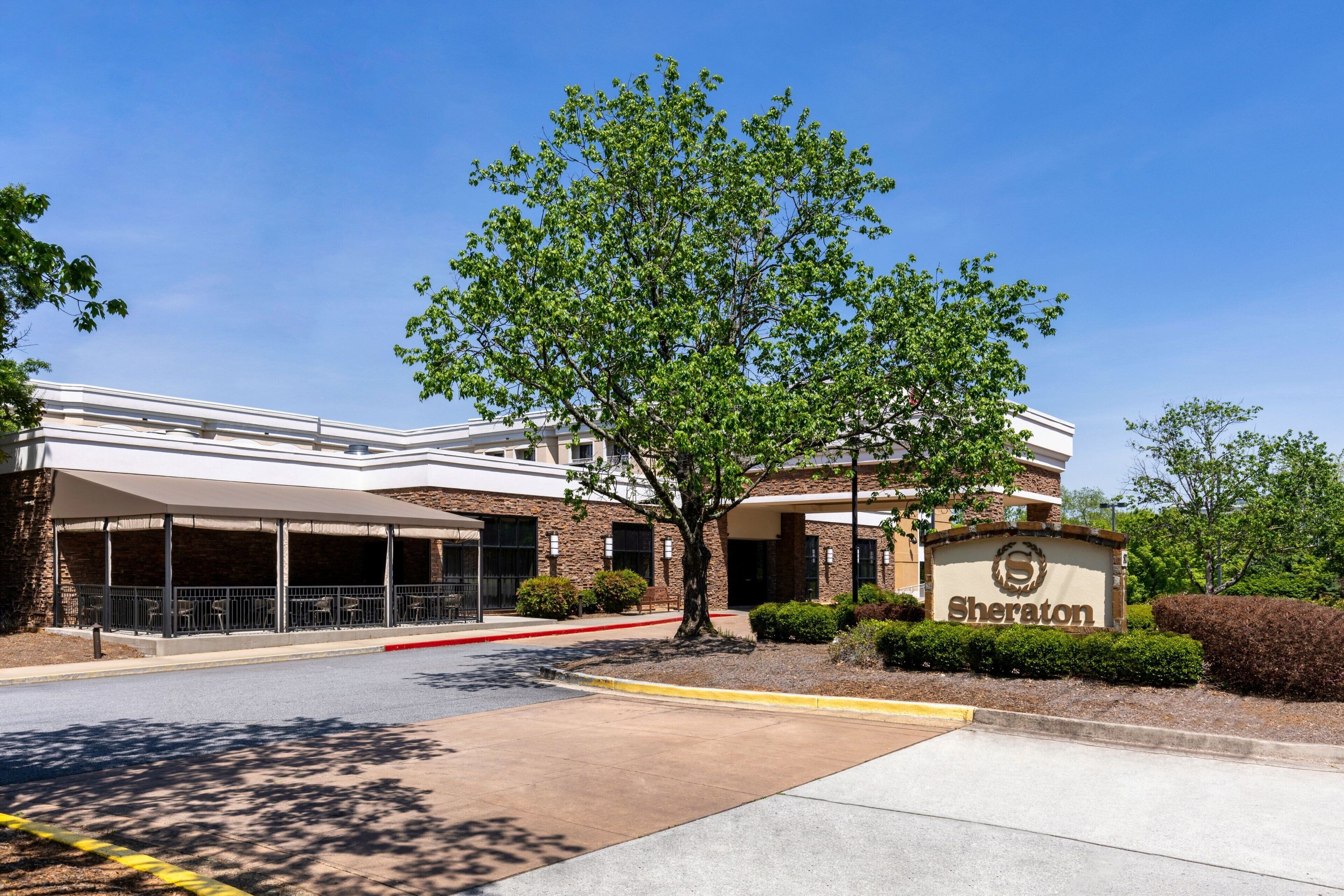 A Sheraton hotel entrance with a covered parking area, a large tree, and a clear blue sky.
