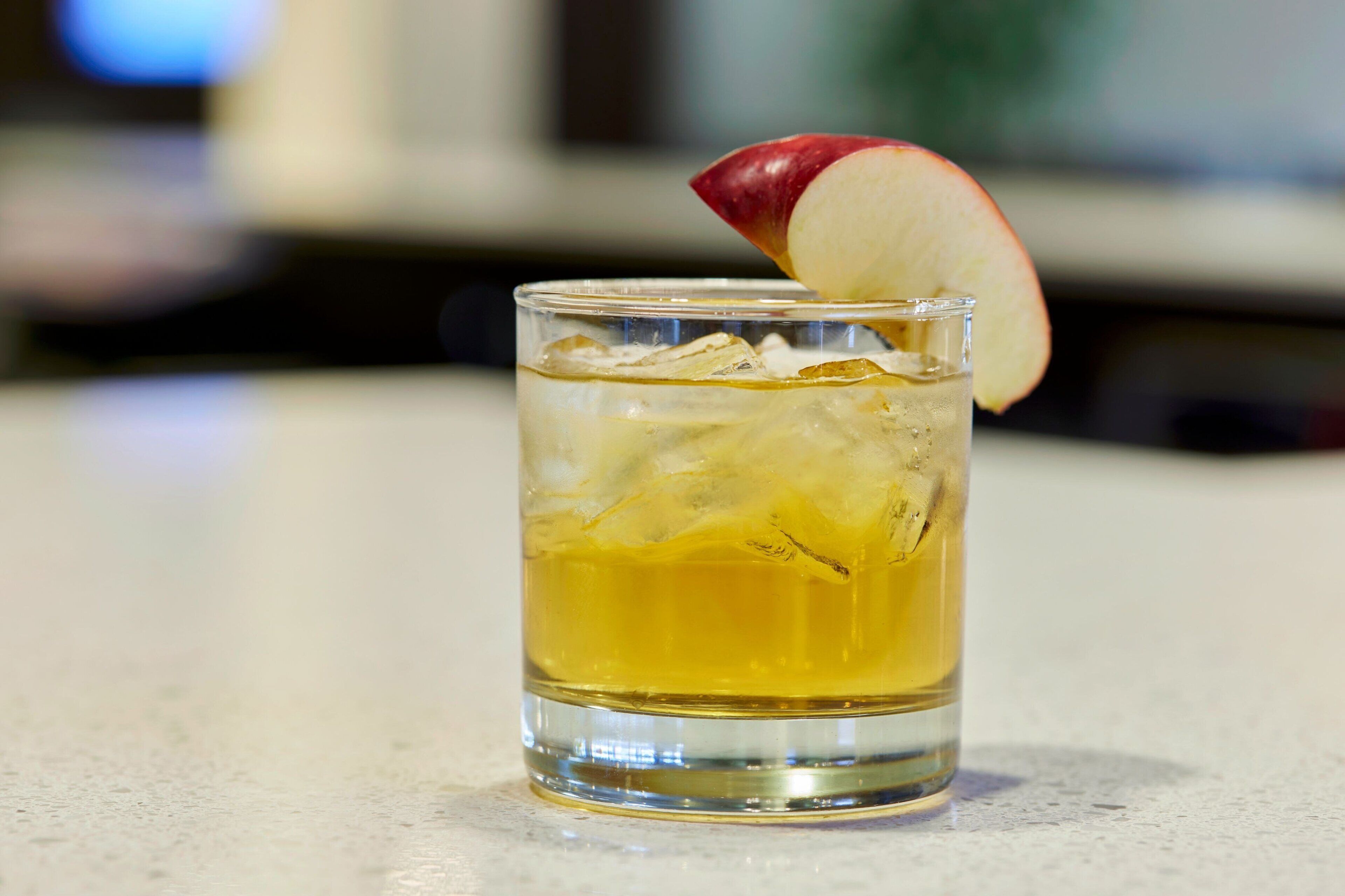 Un verre transparent contenant un liquide jaune et des glaçons, agrémenté d’une tranche de pomme rouge posée sur une surface plane.