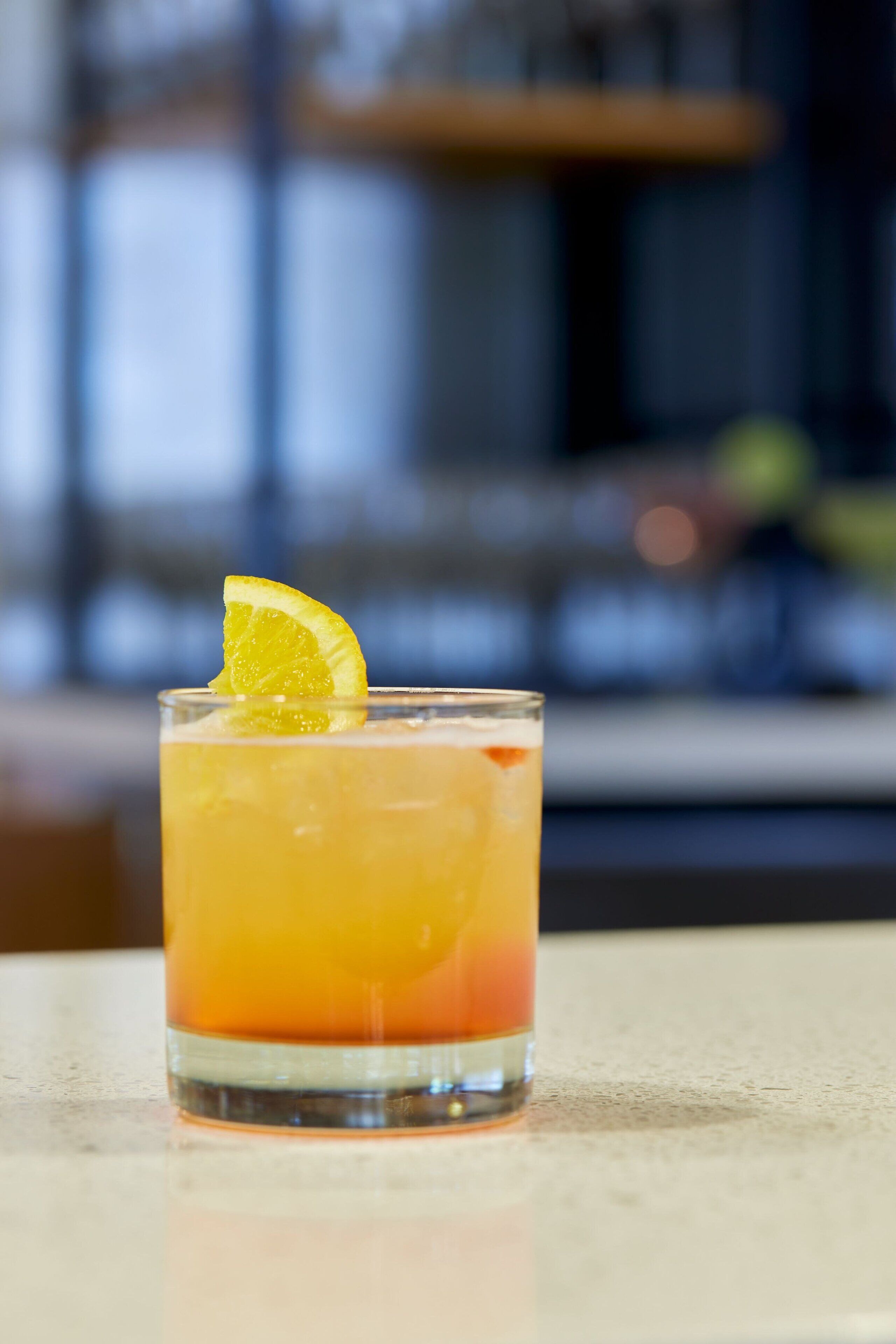 A glass of orange-colored drink with a lemon slice garnish on a table.