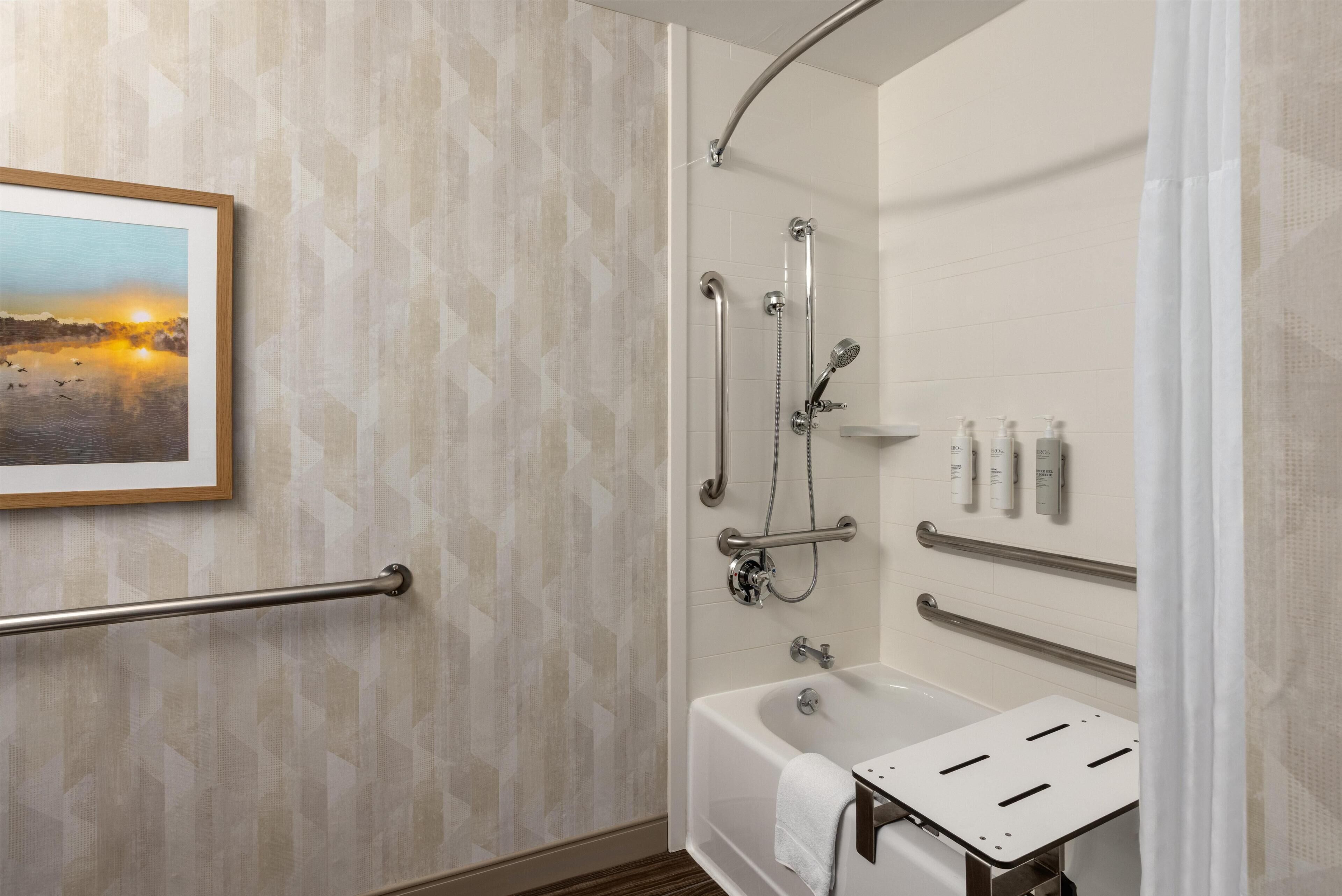 A bathroom with a shower, a bathtub, handrails, and a framed picture on the wall.