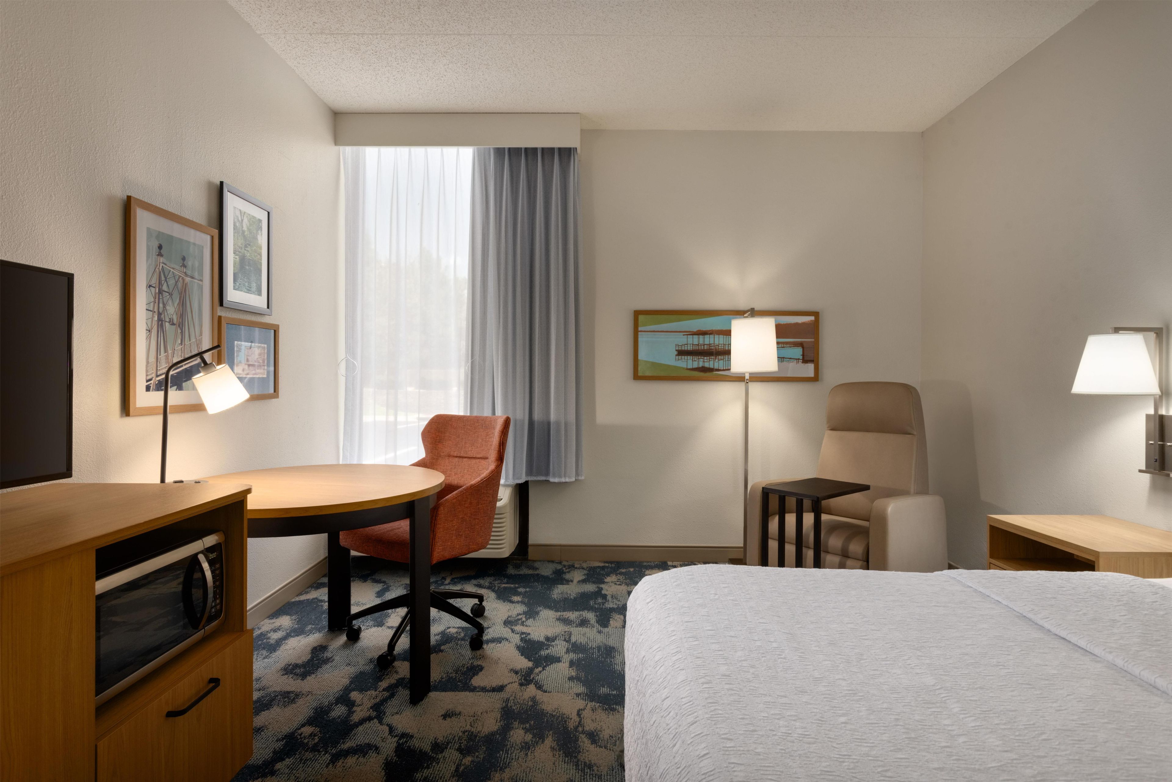 A hotel room with a bed, a desk, a chair, a microwave, a lamp, and a window with curtains.