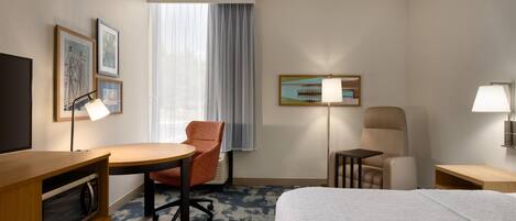 A hotel room with a bed, a desk, a chair, a microwave, a lamp, and a window with curtains.