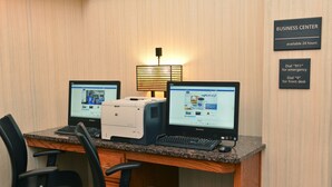 Un centro de negocios equipado con dos computadoras, una impresora y una lámpara. Señalización indica que el centro está disponible las 24 horas y proporciona información de contacto para emergencias y la recepción.