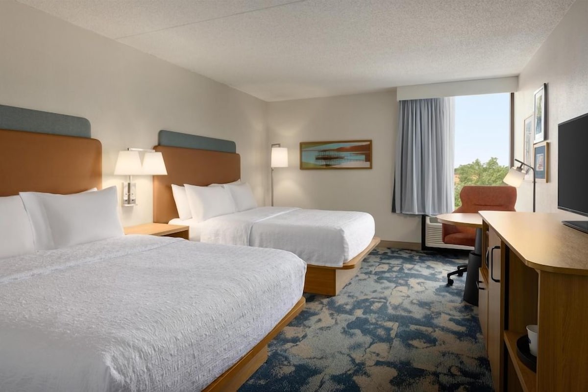 A hotel room with two beds, a desk, a chair, a television, and a window with curtains.