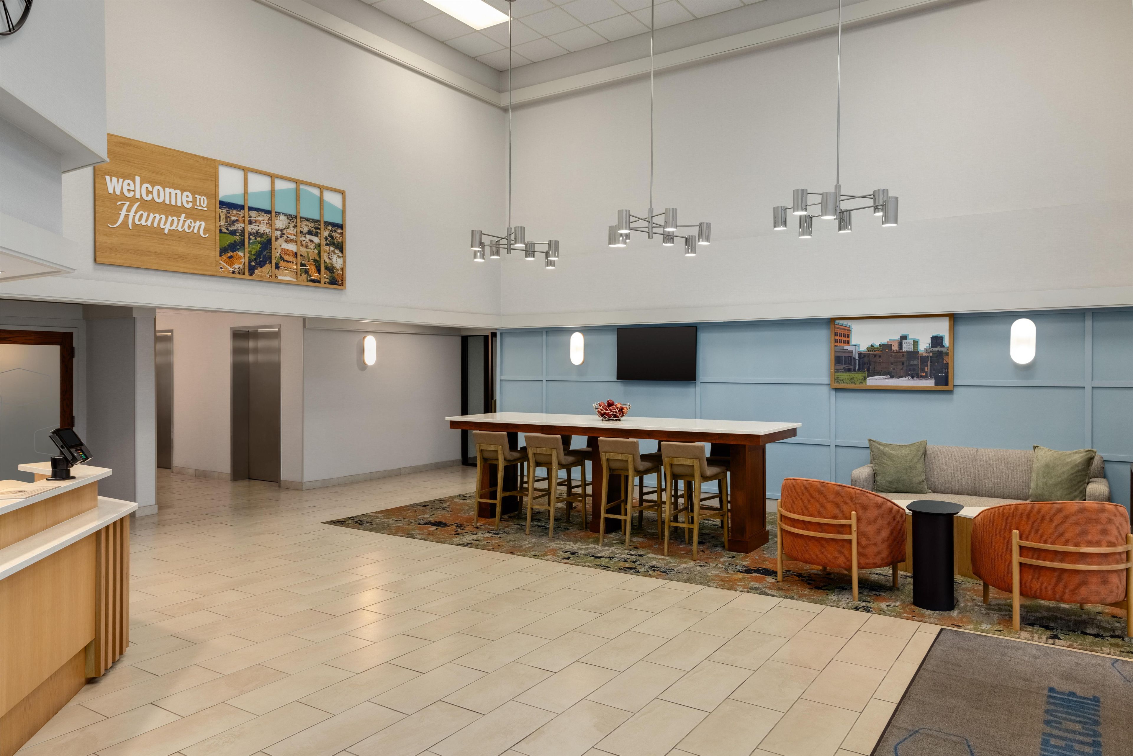 A modern reception area with a 'Welcome to Hampton' sign, a reception desk, a dining area with a table and chairs, a sofa, and a patterned rug.