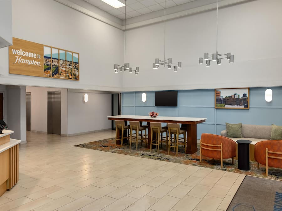 Un'accogliente area di accoglienza moderna con un cartello 'Welcome to Hampton', una reception, un'area ristorazione con tavolo e sedie, un divano e un tappeto decorato.
