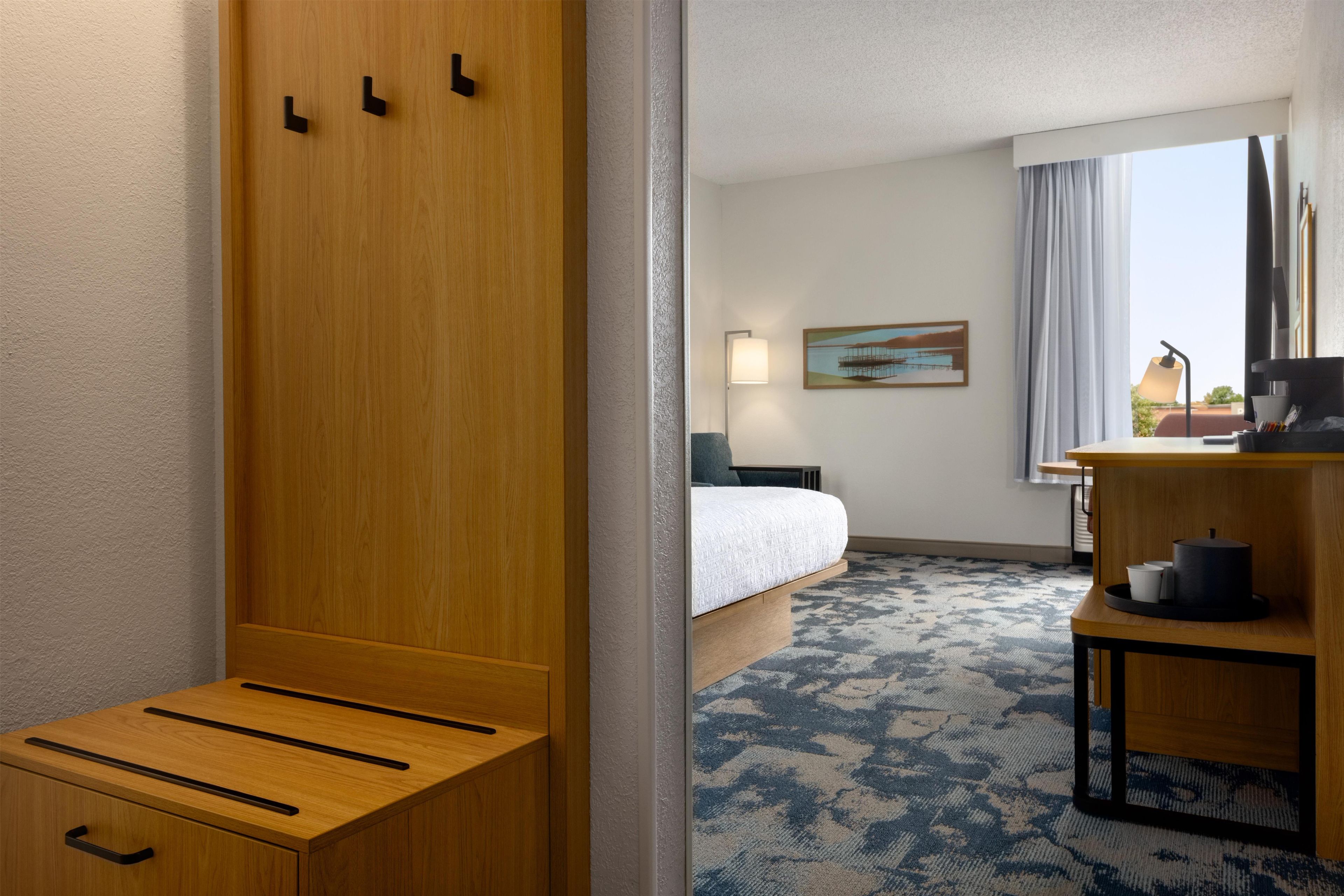 A hotel room with a bed, a desk, a chair, a lamp, a painting, and a view of the outdoors.