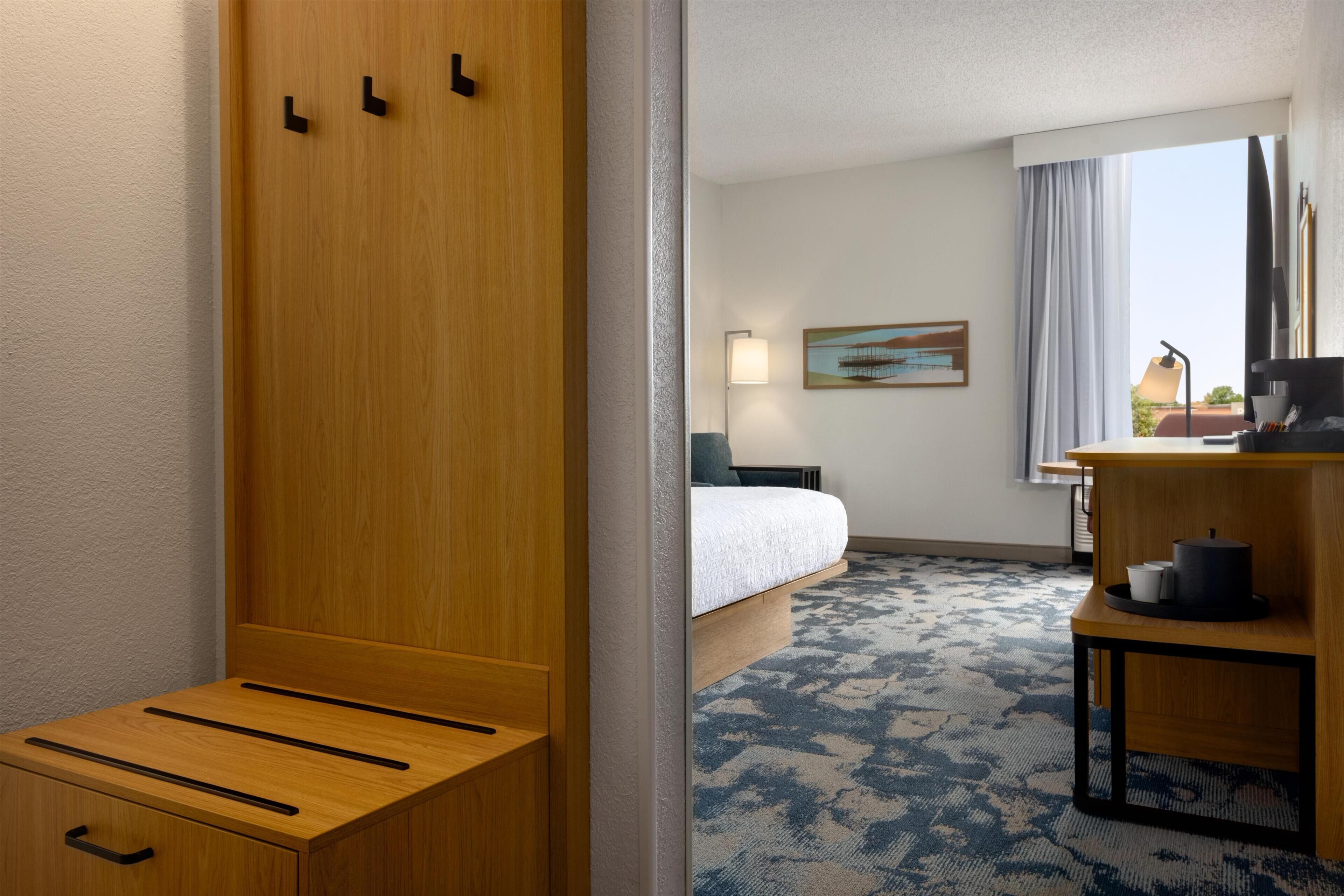 A hotel room with a bed, a desk, a chair, a lamp, a painting, and a view of the outdoors.